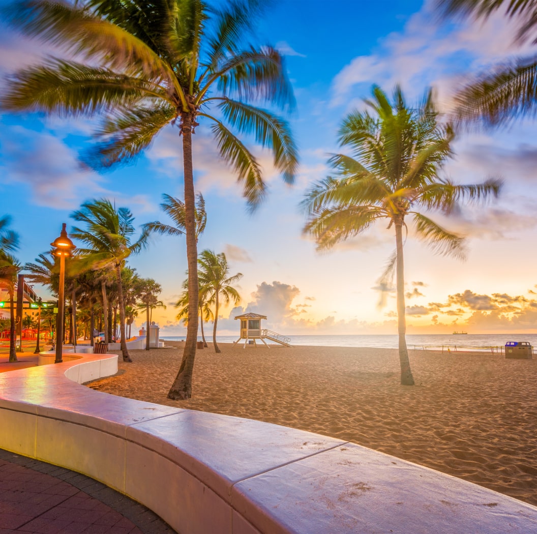Beach scene at sunrise with palm trees, a lifeguard stand, and a curved walkway.