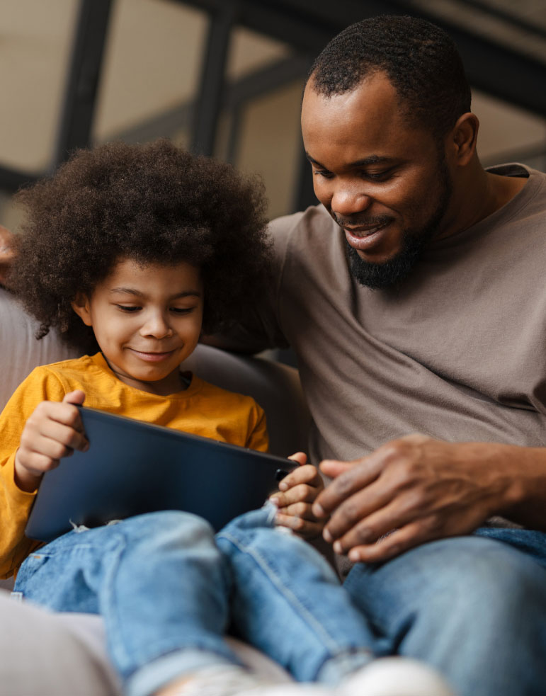 An adult and child sitting on a couch, smiling and looking at a tablet together.