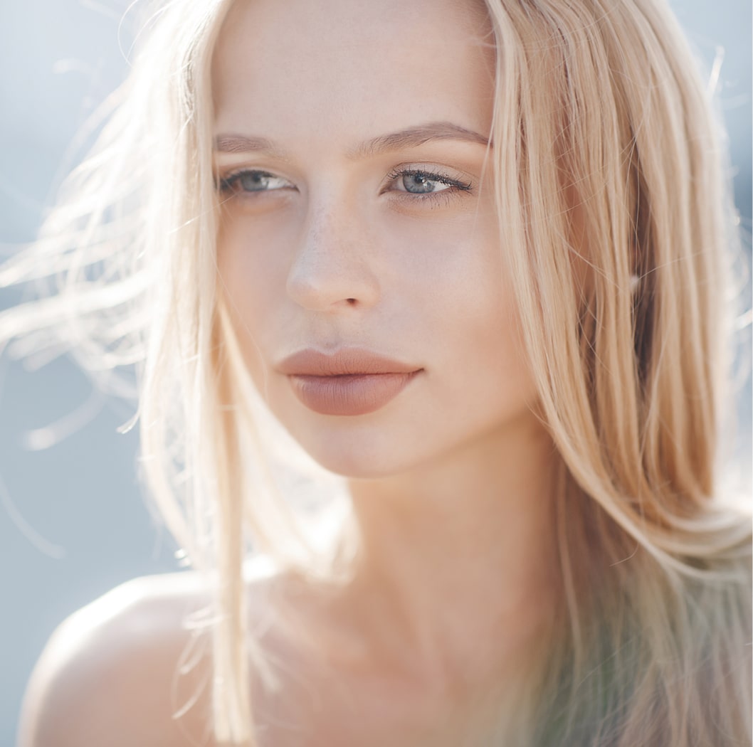 A person with long blonde hair and natural makeup is looking towards the camera in soft lighting.