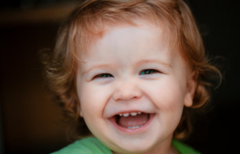 A young child with light brown hair smiles widely, showing small teeth and wearing a green shirt.