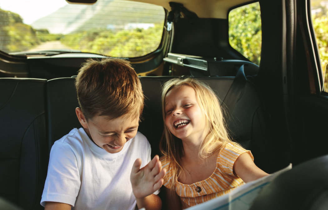 Two young children sit in the backseat of a car, laughing and smiling together during a car ride.