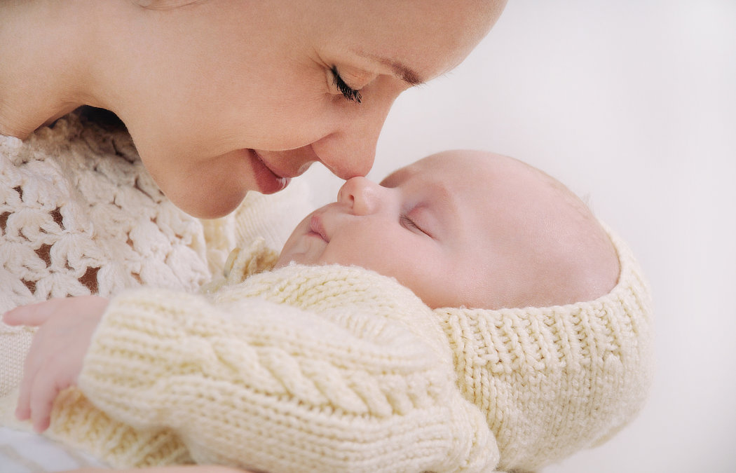 A woman gently touches noses with a sleeping baby dressed in a knitted cream-colored outfit.