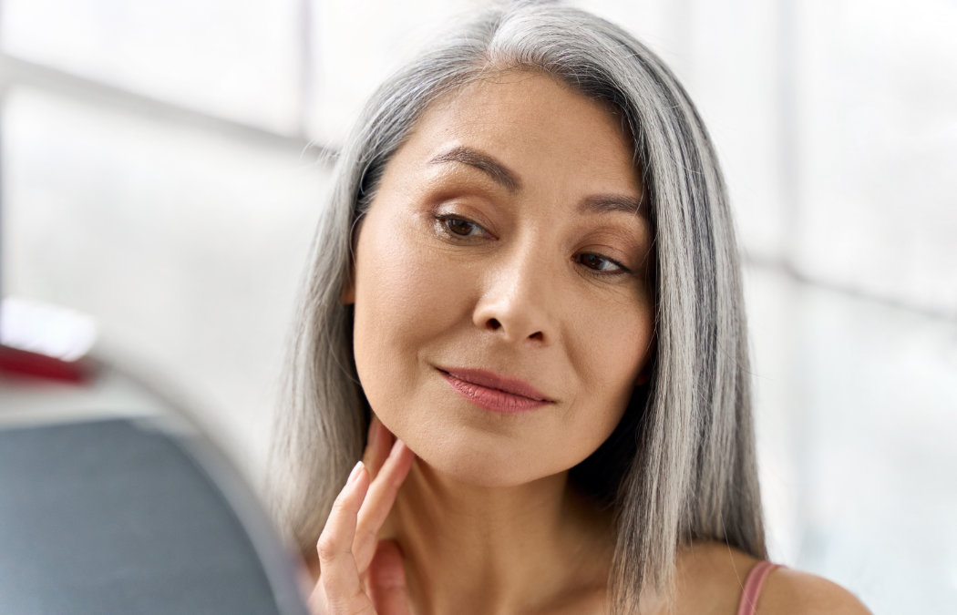 A woman with long gray hair looks at herself in a mirror, touching her neck with her hand.