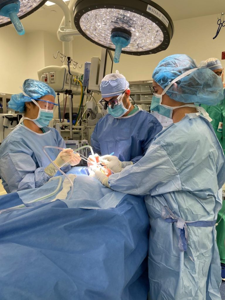 Three surgeons in blue scrubs and masks perform a procedure on a patient in an operating room under bright surgical lights.