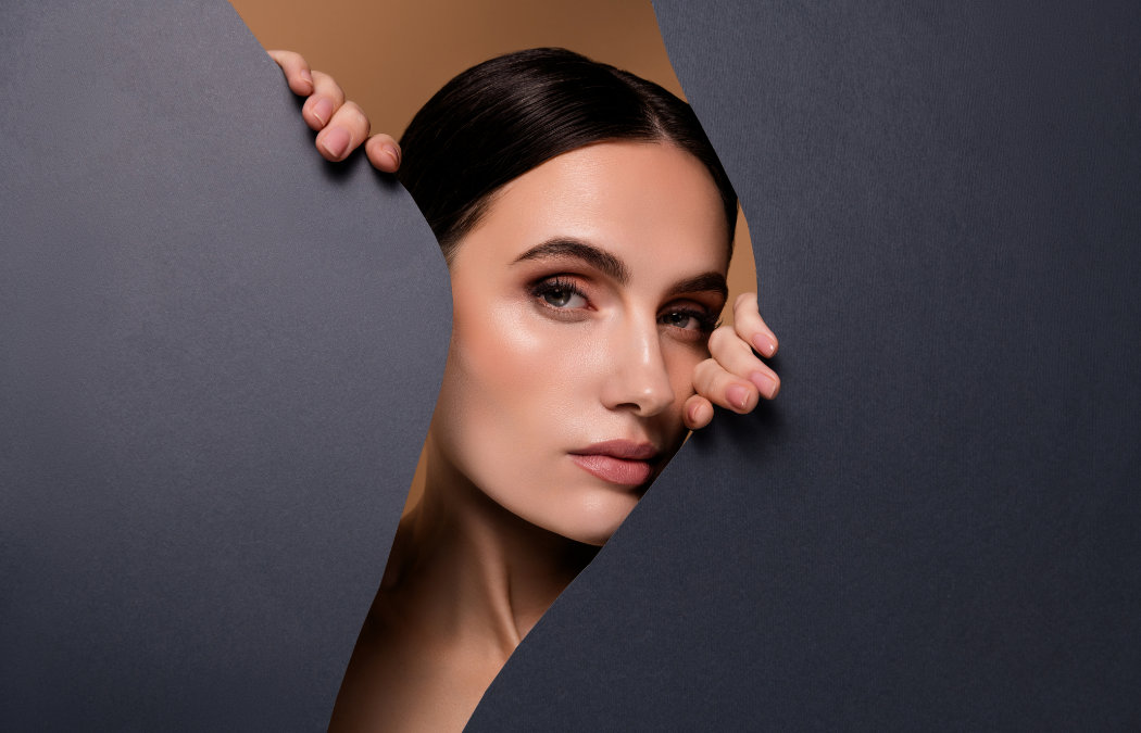 A woman with dark hair and natural makeup peers through a jagged opening in a large dark sheet, looking directly at the camera.