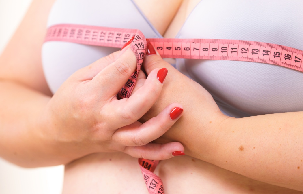 Closeup of female breasts. Plus size fat woman wearing bra using tape measure to check the measurements of her big chest. Bosom, brafitting and underwear concept.