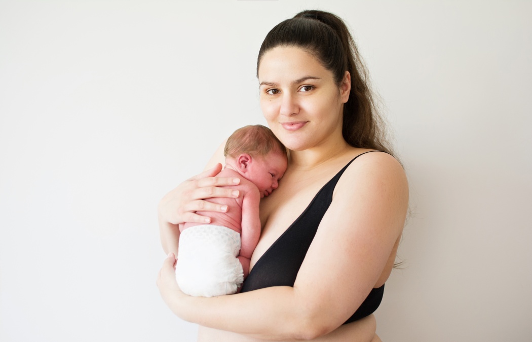 Plus size Caucasian brunette mother smiling and holding her newborn baby. Closeup studio lighting, portrait of mom in black bra and cute baby in a diaper